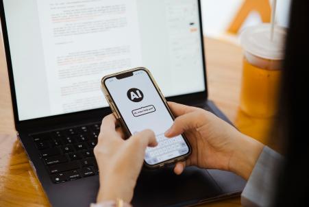 A person uses an AI app on their smartphone while sitting in front of a laptop displaying a cover letter